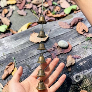 May include: A decorative wind chime with six small, bronze-coloured bells hanging from a chain. The bells are different sizes and are arranged vertically. The background shows wooden planks and fallen leaves.