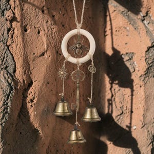 May include: A decorative wind chime featuring a wooden ring with a metal bee in the center. Below the ring hang metal charms, including a sun, key, and bells. The wind chime is suspended by a natural twine cord.