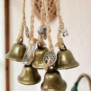 May include: A collection of bronze-colored bells hanging from natural twine. Each bell is adorned with a small charm, including a pentagram, a heart with the word "Bless", a tree of life, a star, and a crescent moon. A small glass bottle with a cork is also included.