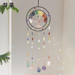 Puede incluir: Un atrapasueños negro con un diseño de árbol de la vida hecho de piedras preciosas de colores. El atrapasueños tiene múltiples hilos de cuentas y piedras preciosas colgando debajo. El anillo superior está decorado con pequeñas cuentas de colores.