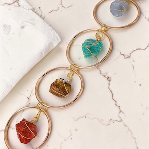 May include: A gold-toned chain necklace featuring four oval links. Each link suspends a different colored gemstone: red, brown, teal, and blue. The gemstones are wrapped in wire. The necklace is displayed on a white and gray marble surface.