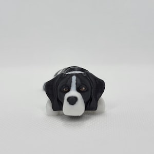 May include: A small, black and white dog figurine. The dog is lying down with its head facing forward. It has brown eyes, a white snout, and a white stripe down the center of its head. The dog's paws are white.