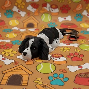 May include: A black and white dog figurine with a keychain attachment rests on a brown background patterned with dog-themed illustrations. The background features paw prints, bones, tennis balls, and dog houses. The dog figurine is approximately 3 inches long.