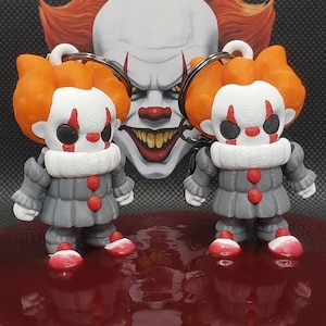 Könnte beinhalten: Zwei Pennywise-Schlüsselanhänger mit orangefarbenem Haar, weißem Gesicht mit roten Markierungen und grauen Outfits mit roten Knöpfen. Die Figuren stehen auf einer glänzenden roten Oberfläche vor einer Pennywise-Illustration mit dem Text HARMAN3D.