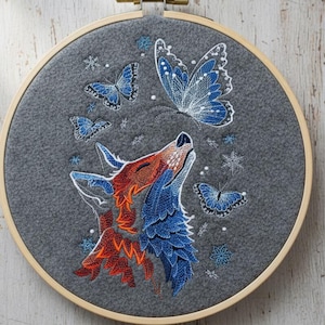 May include: Embroidered hoop art featuring a fox with orange and blue fur, looking up at blue butterflies. The design is on a gray felt background, with snowflakes and a wooden hoop.