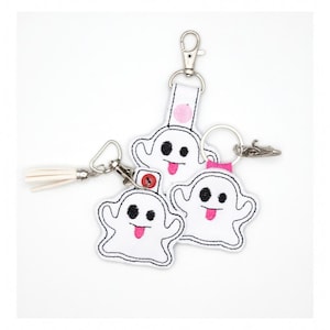 May include: White ghost-shaped keychains with black eyes and pink tongues. Each keychain has a white outline and a metal clasp. One keychain has a white tassel. The keychains are on a white background.