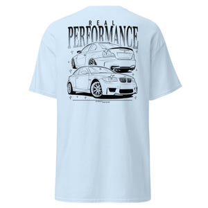 May include: Light blue t-shirt featuring a black and white graphic of two cars with the text "REAL PERFORMANCE" above them. The cars are detailed with outlines, showcasing their design. The shirt is a casual wear item.