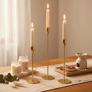 Könnte beinhalten: Drei brennende Kerzen in goldfarbenen Metallhaltern. Die Kerzen sind weiß und verjüngen sich nach oben. Die Halter haben einen kreisförmigen Sockel und einen schlanken Stiel. Die Szene umfasst zwei Kerzengläser und einen kleinen weißen Diffusor.