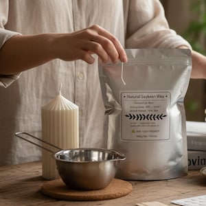 Può includere: Un set per la produzione di candele con una candela bianca alta e scanalata, una pentola in acciaio inossidabile e un sacchetto d'argento di "Natural Saykesan Wax". La borsa ha testo e un disegno a foglie. Su un sottobicchiere di sughero.
