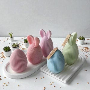 Könnte beinhalten: Osterdekorationen in Pastellfarben. Das Bild zeigt fünf eiförmige und hasenförmige Ornamente. Sie sind auf weißen Tabletts angeordnet, mit kleinen Topfpflanzen im Hintergrund. Die Objekte haben eine gerippte Textur und einige haben Hasenohren.