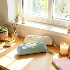 May include: Two semi-cylindrical pencil cases made of smooth material. One case is cream-colored with the name Victoria printed in black script, and the other is light mint green with the name Charlotte printed in black sans-serif font.