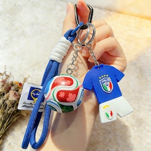 Puede incluir: Un llavero con temática de fútbol con un balón azul y blanco con detalles rojos y un cordón azul. Otro llavero muestra un uniforme de fútbol italiano. Ambos están unidos a un llavero plateado.