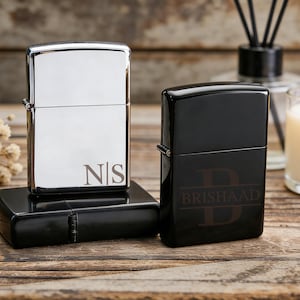 May include: Two personalized lighters. One is silver with the initials "NS", the other is black with the name "BRISHAAD". Both lighters are on a wooden surface, with a candle and diffuser in the background.