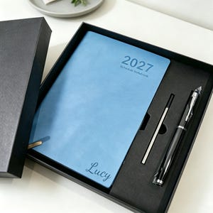 May include: A light blue 2027 schedule notebook with the name Lucy printed on the cover, presented in a black gift box alongside a black pen and a thin metal refill. The notebook cover has a smooth texture and the text 2027 Schedule Notebook.