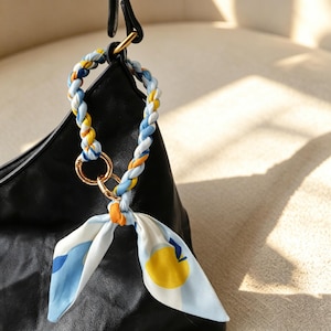 Pode incluir: Um pendente de bolsa trançado em tecido com padrões brancos, amarelos e azuis, com um anel de metal dourado. O acessório está preso a uma bolsa preta e inclui um detalhe de nó em tecido com uma estampa gráfica de limão.