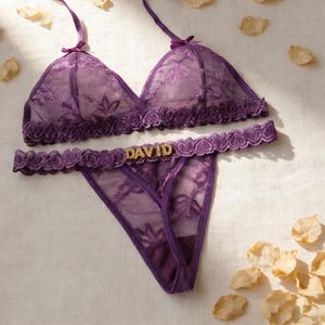 May include: A matching purple lingerie set comprising a bra and thong. The bra has adjustable straps and a delicate floral lace design. The thong features a gold "DAVID" embellishment. The set is made of sheer fabric and trimmed with scalloped edges.