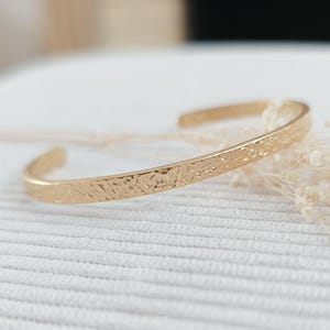Peut inclure: Un bracelet jonc ouvert de couleur or avec une surface texturée. Le bracelet est en métal brillant et présente un design simple et élégant. Le bijou est présenté sur une surface blanche.