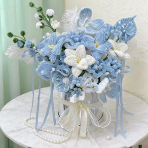 May include: A handmade floral arrangement in a silver container, featuring light blue and white flowers with pearl accents. The bouquet includes ribbons and feathers, creating a delicate display. A pearl necklace is draped nearby.