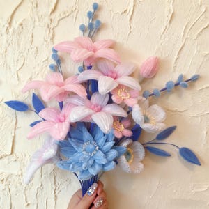 May include: A bouquet of handmade fabric flowers in shades of pink, white, and blue. The arrangement includes lilies, anemones, and a large blue dahlia. The stems are bound together with blue thread, creating a delicate and artistic floral display.