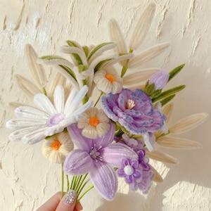 May include: A handcrafted bouquet of fabric flowers in white, purple, and yellow. The bouquet features daisies, lilies, and other floral elements with green stems and leaves. The flowers are held together, creating a vibrant and textured arrangement.