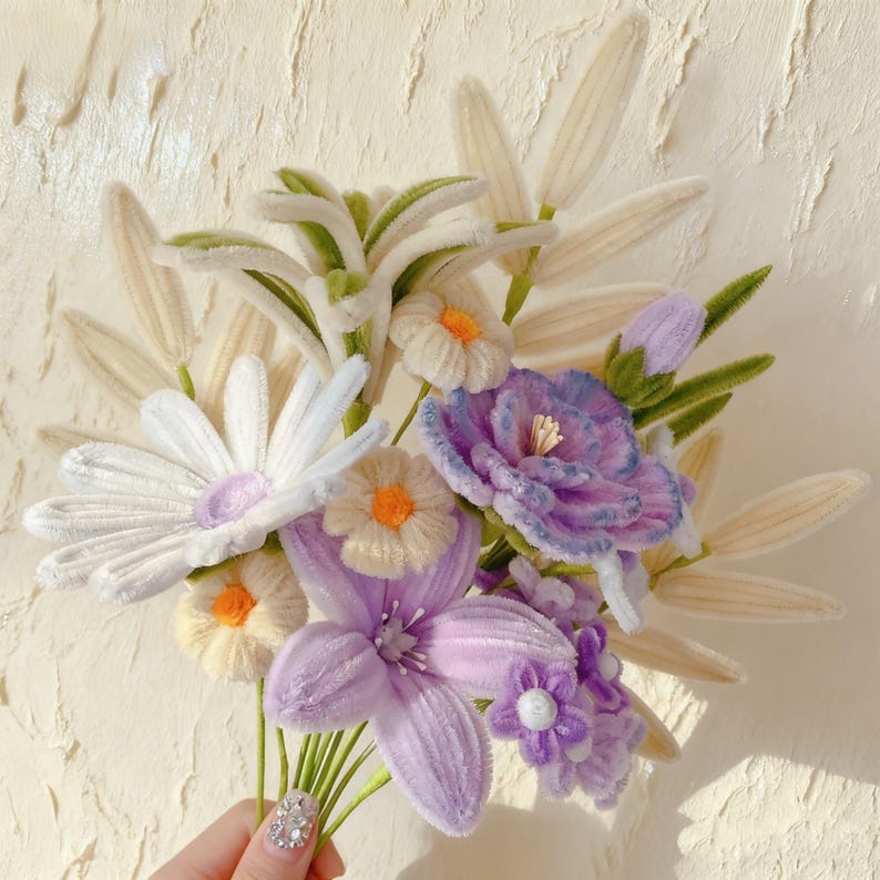 May include: A handmade bouquet of fabric flowers in shades of white, purple, and yellow. The bouquet includes daisies, lilies, and other floral elements with green stems and leaves. The flowers are held together, creating a vibrant and textured arrangement.