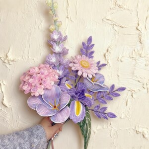 May include: A bouquet of handmade paper flowers in shades of purple, pink, and yellow. The arrangement includes irises, daisies, and other floral elements. The flowers are held against a textured, off-white background.