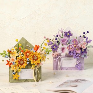 May include: Two decorative floral arrangements in the shape of envelopes. One is green and filled with yellow, orange, and white flowers. The other is purple and filled with purple and pink flowers. Both have small cards and pearl accents.