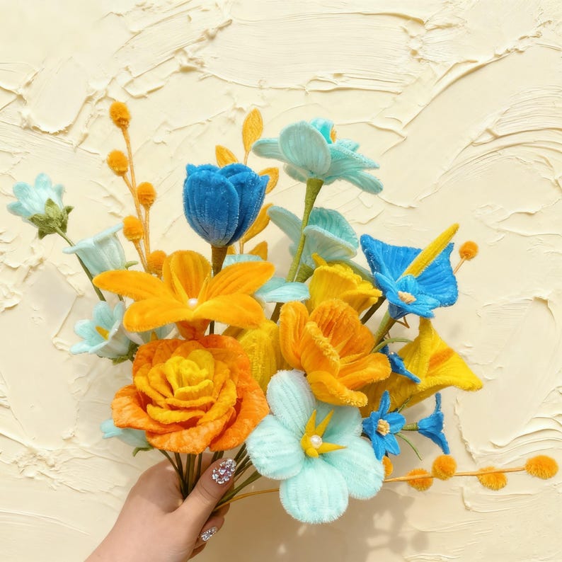 May include: A bouquet of handmade, colorful flowers in shades of blue, yellow, and orange. The flowers are made of a soft, textured material and arranged against a textured, cream-colored background. The bouquet includes roses, tulips, and other floral varieties.