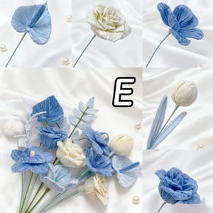 May include: A collection of handmade fabric flowers in shades of blue and white. The arrangement includes various flower types, such as roses, tulips, and anthuriums, with green stems. The flowers are arranged on a white satin background with scattered pearls.