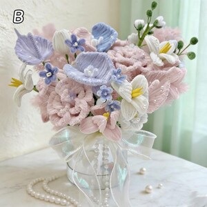 May include: A fabric flower bouquet in a small, round, silver container. The arrangement includes pink, white, and blue flowers with pearl accents. An iridescent ribbon and a pearl necklace decorate the container.