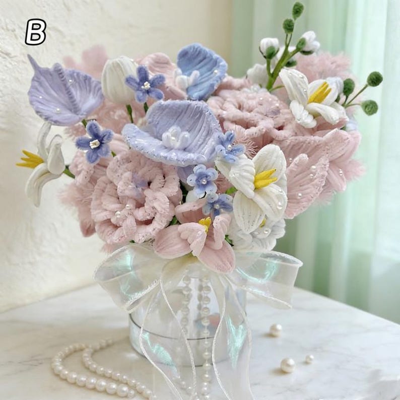 May include: A fabric flower bouquet in a small, round, silver container. The arrangement includes pink, white, and blue flowers with pearl accents. An iridescent ribbon and a pearl necklace decorate the container.