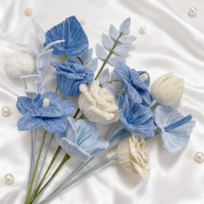 May include: A bouquet of artificial flowers in shades of blue and cream. The arrangement includes roses, tulips, and other floral elements with green stems. The flowers are arranged on a white satin background with scattered pearls.