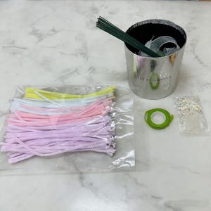 May include: A floral arrangement kit with various supplies. Includes a silver bucket with ribbon and green floral wire, a package of pastel-coloured pipe cleaners, green floral tape, and small silver embellishments.