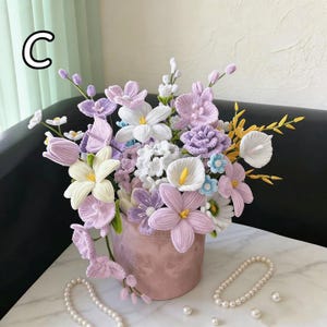 May include: A crochet floral display in a pink vase, showcasing lavender, white, and yellow flowers. The arrangement includes calla lilies, tulips, and other detailed blooms. The letter "C" is visible in the upper left corner. The display is on a marble surface.