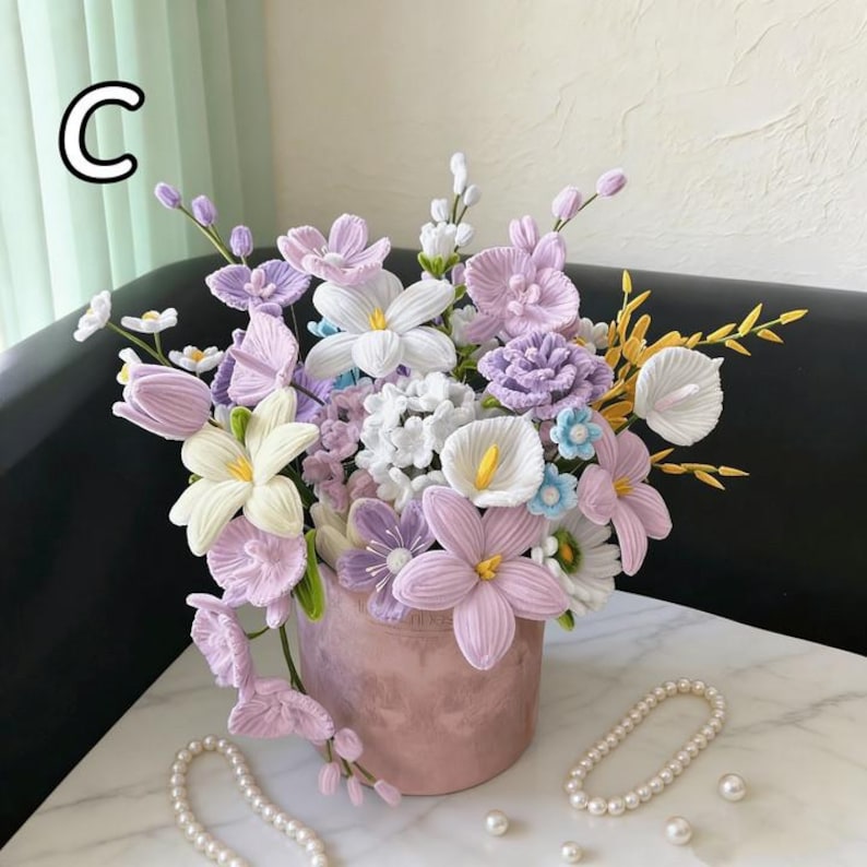 May include: A crochet floral display in a pink vase, showcasing lavender, white, and yellow flowers. The arrangement includes calla lilies, tulips, and other detailed blooms. The letter "C" is visible in the upper left corner. The display is on a marble surface.