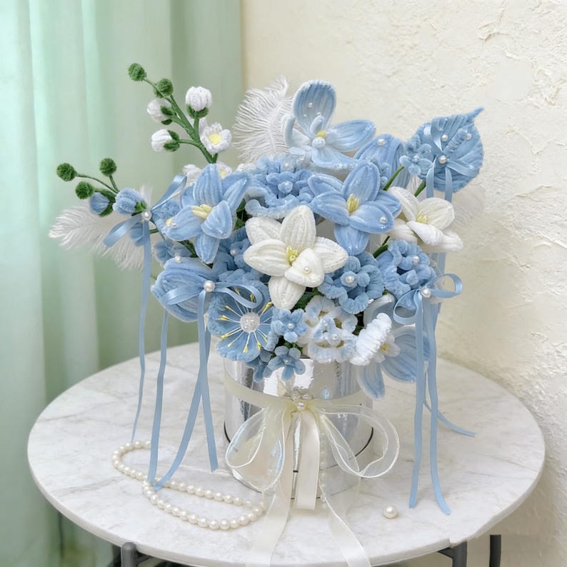 May include: A handmade floral arrangement in a silver container, featuring light blue and white flowers with pearl accents. The bouquet includes ribbons and feathers, creating a delicate display. A pearl necklace is draped nearby.