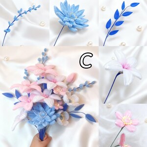 May include: A collection of handmade fabric flowers in various styles and colors. The assortment includes blue, pink, and white blossoms, some with beaded centers. The flowers are arranged on stems and displayed against a white satin background with scattered pearls.
