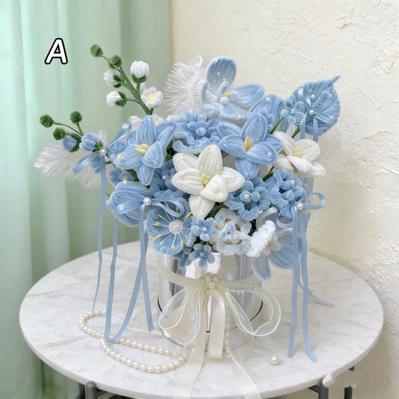 May include: A handmade floral arrangement in a silver container, featuring light blue and white fabric flowers. The bouquet includes ribbons, pearls, and feathers, creating a delicate and elegant aesthetic. The arrangement is displayed on a white marble table.
