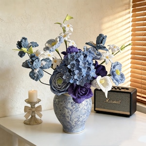 May include: A floral arrangement in a blue and white vase, featuring blue, white, and purple flowers. The arrangement includes hydrangeas, irises, and other blooms. A small Marshall speaker and a candle holder with a lit candle are also present.