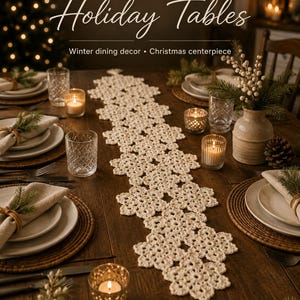 Peut inclure: Un chemin de table en crochet de couleur crème présentant un motif de flocons de neige répétitif avec un pompon à chaque extrémité. Le chemin est disposé sur une table en bois avec des assiettes, des verres, des bougies et du feuillage.