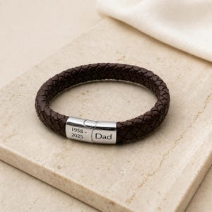 May include: A dark brown braided leather bracelet featuring a polished silver-tone metal clasp. The clasp is engraved with the text 1958 - 2025 on one side and the word Dad on the other side. The bracelet has a circular shape and a woven texture.