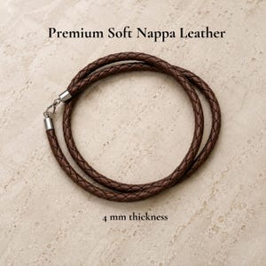 Può includere: Una collana in corda di pelle intrecciata marrone scuro con chiusura a moschettone e terminali in metallo argentato. Il testo Premium Soft Nappa Leather e 4 mm thickness è visibile. La corda è disposta in un cerchio con uno spessore di 4 mm.