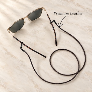 May include: A pair of sunglasses with dark lenses and a tortoiseshell frame attached to a braided dark brown cord. The cord is labeled with the text Premium Leather and features black connectors that secure the strap to the temple arms.