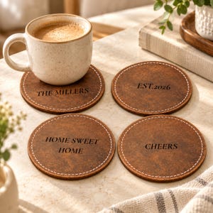 May include: Four round brown coasters with white stitched edges. Each coaster features black text: THE MILLERS, EST. 2026, HOME SWEET HOME, and CHEERS. A speckled beige mug filled with coffee sits on the coaster labeled THE MILLERS.