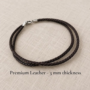 May include: A dark brown braided leather cord necklace or bracelet featuring a silver-toned metal clasp. The cord has a circular shape and is labeled with the text Premium Leather - 3 mm thickness, indicating its material and diameter.