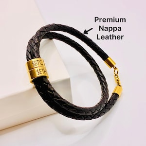 Può includere: Un braccialetto in pelle intrecciata nera con dettagli dorati. Il braccialetto presenta due fili di pelle intrecciata e una chiusura dorata. Il braccialetto ha un cilindro dorato con il testo "$BTC" e "$ETH". È visibile anche il testo "Premium Nappa Leather".