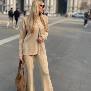 May include: A beige knit two-piece set consisting of a long-sleeved cardigan with a ribbed hem and matching flared trousers with a center seam. The outfit is paired with a large brown suede hobo bag featuring fringe details and dark sunglasses.