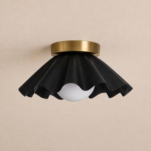 May include: A flush mount ceiling light fixture featuring a matte black, ruffled, wide-brimmed metal shade attached to a cylindrical brushed brass base, with a single spherical white glass light bulb centered within the shade's opening.