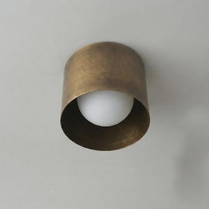 Op de afbeelding: Een cilindrische plafondlamp met een geborstelde antiek messing afwerking op de buitenste metalen behuizing en een enkele bolvormige witte glazen lamp gecentreerd in de open ronde opening van de metalen cilinder.