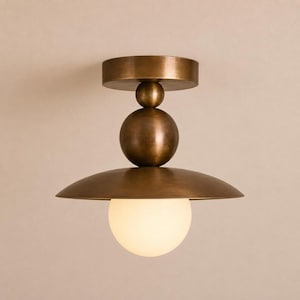 May include: A flush mount ceiling light fixture featuring a circular antique brass canopy, a central vertical stem with two stacked spheres, a wide flat disc-shaped shade, and a single frosted white glass globe bulb at the bottom center.